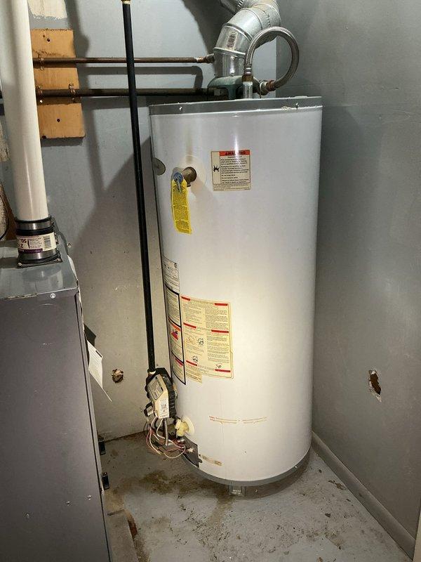 Conducted water heater replacement estimate for residential property. Assessed existing standard white cylindrical tank-type water heater located in basement utility area with visible age-related wear and discoloration. Evaluated current installation including top-mounted exhaust vent with 90-degree metallic ductwork, copper and PVC plumbing connections, gas line with yellow-handled valve, and electrical wiring. Documented equipment specifications, surrounding HVAC infrastructure, and utility room configuration through photographic documentation to provide accurate replacement proposal.