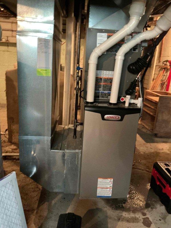 Responded to service call for moisture accumulation under furnace during air conditioning operation. Upon inspection, found condensate drainage issue from central air system affecting Lennox furnace installation. Examined PVC condensate drain lines, evaporator coil drainage, and surrounding equipment for proper operation. Verified condensate pump function, cleared any blockages in drain lines, and ensured proper pitch and routing of white PVC drainage piping extending from unit. Inspected furnace base and surrounding floor area for moisture damage and confirmed source of water accumulation. Checked air handler and ductwork connections for secondary drain pan overflow or leakage. Tested system operation with air conditioning running to verify proper condensate removal and confirmed no further moisture issues at furnace base upon completion.