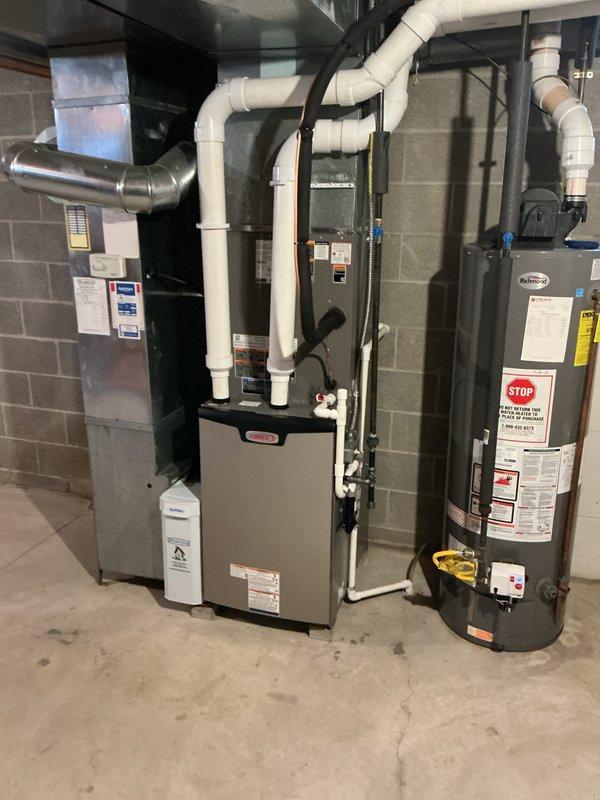 # HVAC Install - Complete System

Completed installation of a 90% efficiency furnace system with integrated HVAC components in mechanical room. Installed new beige cabinet furnace unit as primary heating source, positioned centrally in equipment lineup. Mounted metallic air handler with connecting ductwork for proper air distribution throughout structure. Configured white PVC venting system extending from furnace top for combustion air intake and exhaust requirements per manufacturer specifications.

Installed water heater unit with appropriate safety labeling and warning placards clearly visible on front panel. Established all necessary gas line connections using black pipe with blue-handled shutoff valve for system isolation capability. Installed brass fittings at connection points to ensure secure, leak-free operation. Routed plumbing connections between units for integrated system performance.

Completed ductwork installation including foam-insulated supply ducts and metallic flexi