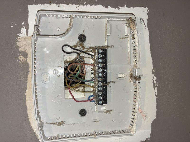 Responded to no cooling call and inspected thermostat at 120 service call location. Found significant corrosion on thermostat wiring terminals and connections, indicating moisture intrusion or age-related deterioration. Documented existing wiring configuration with multiple low-voltage connections (R, Y, G, B, O, C) before removal, noting rust-colored oxidation on wire bundle and terminal block that was preventing proper electrical contact and system communication.
