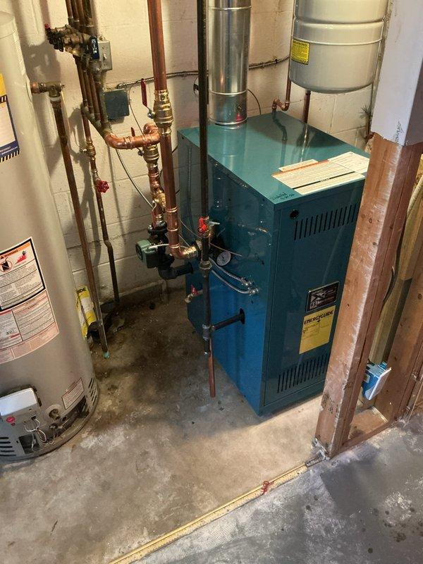 Successfully installed a new hot water boiler system in the basement mechanical room. The installation included mounting the teal-colored boiler unit, connecting extensive copper piping with multiple valves and fittings, installing a gray expansion tank on top of the unit, and routing the metallic flue pipe for proper venting. All plumbing connections were completed with brass valves and red-handled shut-offs integrated into the system alongside the existing water heater. The boiler was positioned against the concrete block wall with proper clearances maintained, all piping secured and supported according to code requirements, and the system prepared for startup and operation.