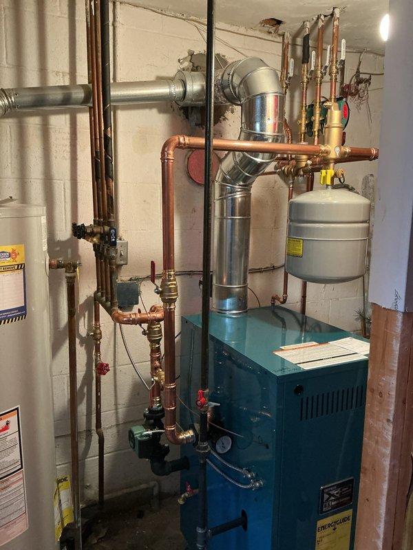 Successfully installed a new hot water boiler system in the basement mechanical room. The installation included mounting the teal-colored boiler unit, connecting extensive copper piping with multiple valves and fittings, installing a gray expansion tank on top of the unit, and routing the metallic flue pipe for proper venting. All plumbing connections were completed with brass valves and red-handled shut-offs integrated into the system alongside the existing water heater. The boiler was positioned against the concrete block wall with proper clearances maintained, all piping secured and supported according to code requirements, and the system prepared for startup and operation.