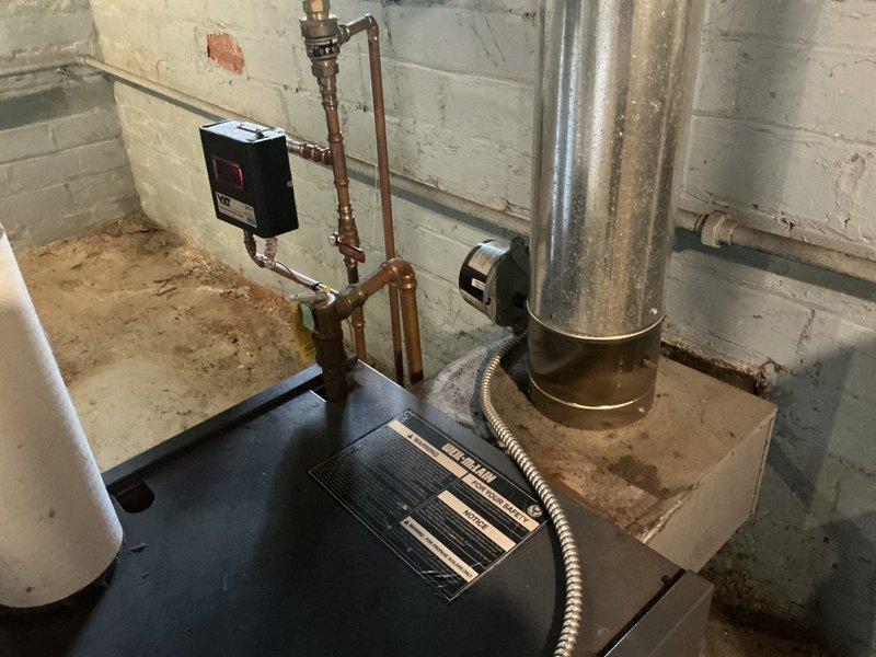 Conducted on-site evaluation of existing steam boiler system in basement utility area. Assessed condition of aging copper piping network showing significant corrosion and patina buildup, galvanized metal flue pipe with surface discoloration and rust spots, and digital control monitoring unit currently displaying operational status. Documented system components including vertical and horizontal pipe runs, fittings, valves, wall-mounted control boxes, and flexible metal conduit connections for estimate preparation.