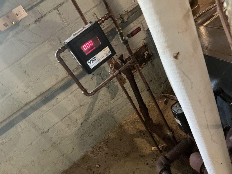 Conducted on-site evaluation of existing steam boiler system in basement utility area. Assessed condition of aging copper piping network showing significant corrosion and patina buildup, galvanized metal flue pipe with surface discoloration and rust spots, and digital control monitoring unit currently displaying operational status. Documented system components including vertical and horizontal pipe runs, fittings, valves, wall-mounted control boxes, and flexible metal conduit connections for estimate preparation.