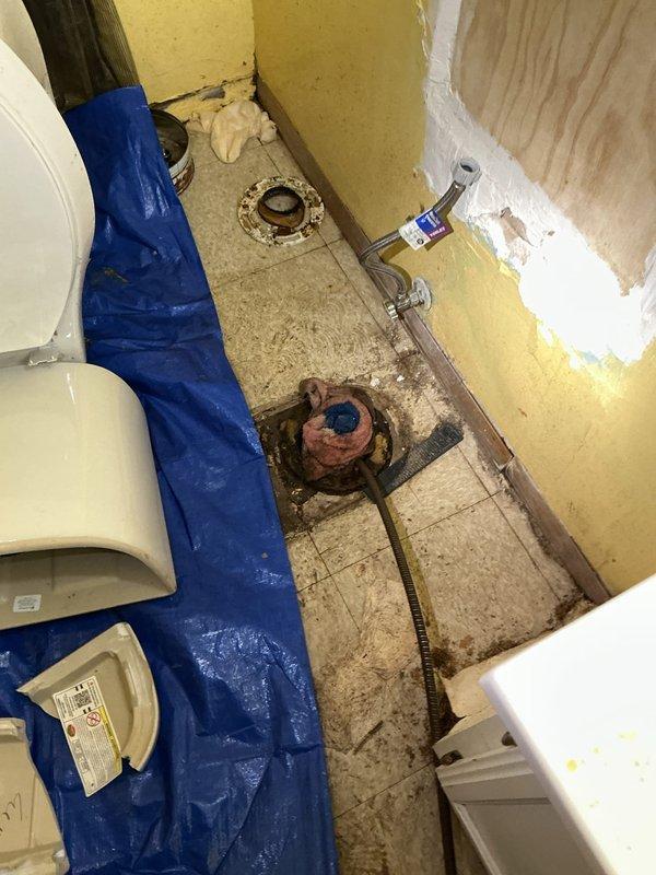 Performed video camera inspection of main drain line to assess condition and identify any blockages or structural issues. Documentation captured showing bathroom floor area with exposed plumbing components including floor drains and flanges, along with utility space drain location where camera access was established for thorough line evaluation.