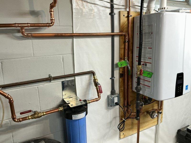 Completed tankless water heater installation in residential utility room, including full system integration with existing copper supply lines, gas line connections, and whole-house filtration system mounted on wooden backing board against concrete block wall. Installed exterior PVC condensate drainage system with proper venting and cleanout access on building exterior, secured to siding with appropriate mounting hardware and terminating above grade level in mulched area.