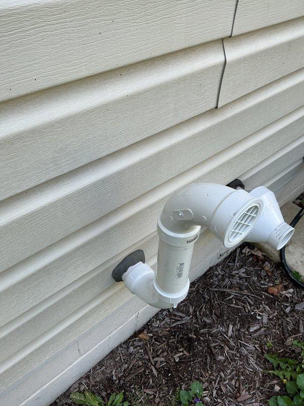 Completed tankless water heater installation in residential utility room, including full system integration with existing copper supply lines, gas line connections, and whole-house filtration system mounted on wooden backing board against concrete block wall. Installed exterior PVC condensate drainage system with proper venting and cleanout access on building exterior, secured to siding with appropriate mounting hardware and terminating above grade level in mulched area.