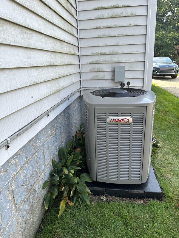 Completed full HVAC system installation of an 80,000 BTU Lennox air conditioning unit at residential property. Mounted new condenser on black composite equipment pad against exterior foundation wall with proper clearance for airflow and service access. Installed gray electrical conduit along exterior wall connecting to weatherproof disconnect box with dual 20A Littelfuse fuses mounted in approved fuse holder assembly. Verified all electrical connections meet code requirements and unit is properly grounded. System tested and confirmed operational with adequate refrigerant charge and airflow. All components functioning within manufacturer specifications.