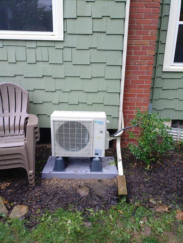 Completed mini split system installation with outdoor condensing unit mounted on concrete pad against brick chimney. Installation required extension ladder for elevated work and included running refrigerant lines along exterior wall. Discovered and replaced failed electrical disconnect with severe corrosion and heat damage on fuse holders, ensuring safe electrical connection for new Daikin unit.