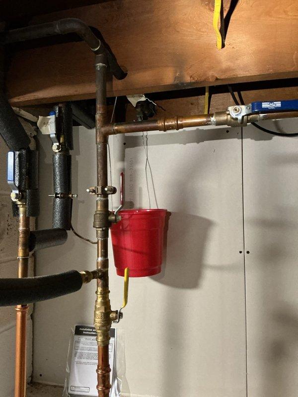 Responded to service call for water heater leaking approximately half cup of water daily. Upon inspection, identified leak originating from pressure relief valve assembly. Photographed existing plumbing configuration showing copper supply lines, brass fittings, and T&P relief valve with discharge tube. Placed collection bucket beneath leak point to contain water discharge. Documented condition of water heater connections, shutoff valves, and surrounding plumbing infrastructure for repair assessment and customer records.