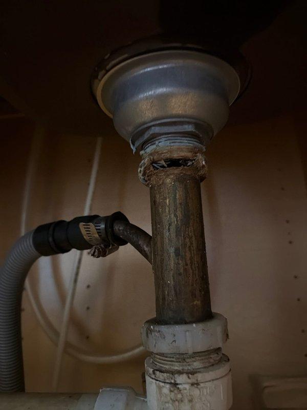 Responded to kitchen double sink drain leak causing water puddles on floor. Upon inspection, found severely corroded and deteriorated drain tailpiece assembly with significant surface degradation and discoloration. The vertical drain pipe section showed extensive weathering and deterioration at connection points, causing the leak between the sink drain and P-trap assembly. Replaced the failed drain tailpiece and associated fittings with new components. Reconnected dishwasher drain line to new assembly. Tested system for proper drainage and verified all connections were leak-free before completing service.