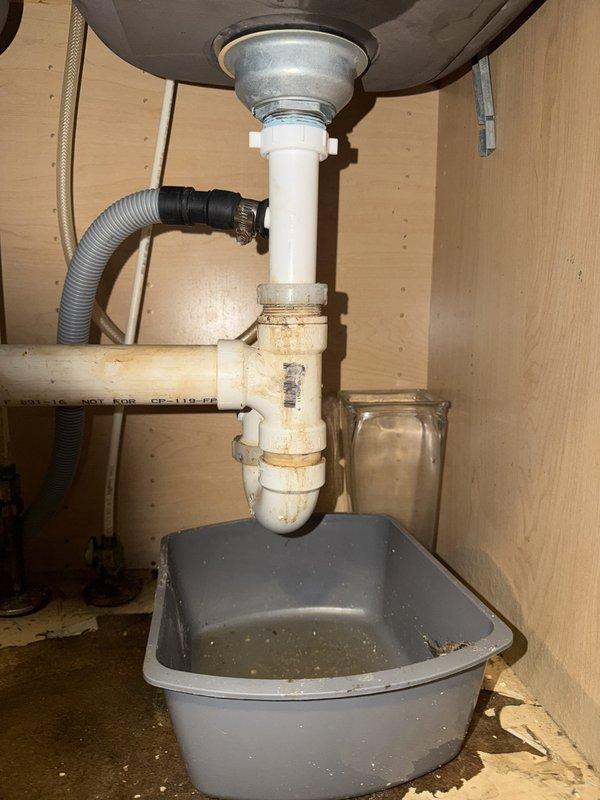 Responded to kitchen double sink drain leak causing water puddles on floor. Upon inspection, found severely corroded and deteriorated drain tailpiece assembly with significant surface degradation and discoloration. The vertical drain pipe section showed extensive weathering and deterioration at connection points, causing the leak between the sink drain and P-trap assembly. Replaced the failed drain tailpiece and associated fittings with new components. Reconnected dishwasher drain line to new assembly. Tested system for proper drainage and verified all connections were leak-free before completing service.