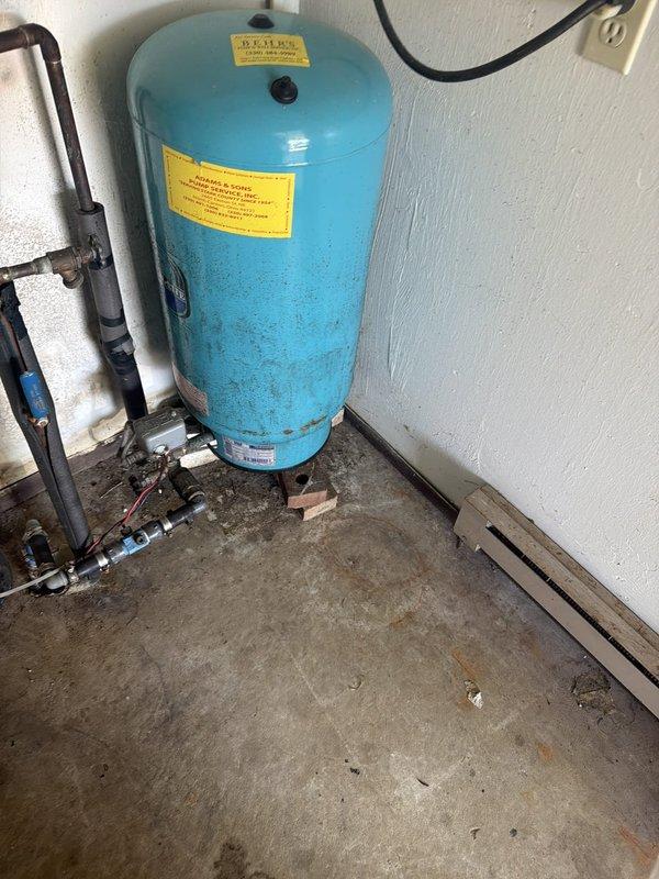 Responded to a complaint of odorous water and inspected the well system, including the pressure tank, water treatment tanks with control valve, and associated piping, to determine whether the source was the water heater or the well supply. Observed a weathered, corroded pressure tank, soiled treatment tanks, and mildew growth in the utility space, and documented all conditions with photos. Findings suggest the odor is likely related to the well water and treatment system rather than the water heater, with further testing and servicing of the treatment equipment recommended.