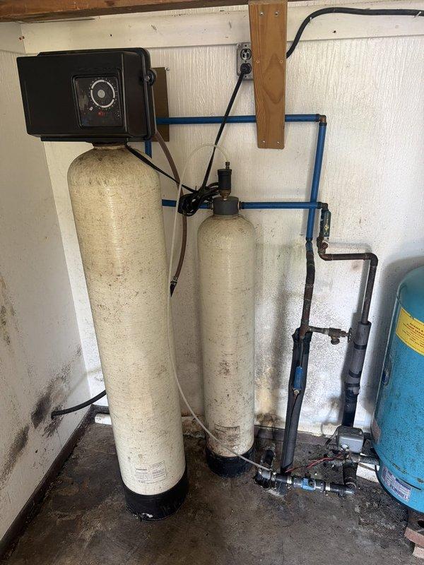 Responded to a complaint of odorous water and inspected the well system, including the pressure tank, water treatment tanks with control valve, and associated piping, to determine whether the source was the water heater or the well supply. Observed a weathered, corroded pressure tank, soiled treatment tanks, and mildew growth in the utility space, and documented all conditions with photos. Findings suggest the odor is likely related to the well water and treatment system rather than the water heater, with further testing and servicing of the treatment equipment recommended.