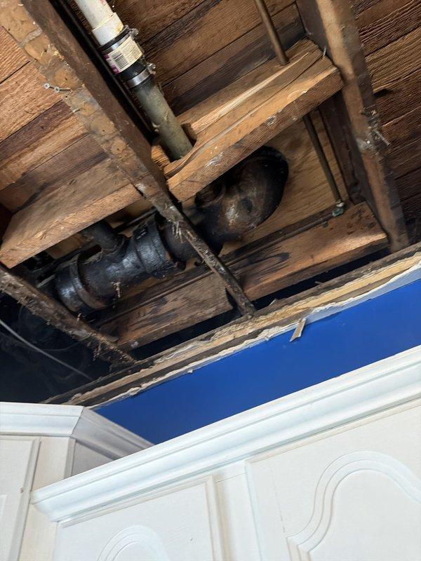 Inspected the open kitchen ceiling cavity to trace an intermittent leak and found moisture-stained framing around the cast iron drain assembly beneath the upstairs toilet, including the banded coupling and elbow transitions to the PVC drain line. Repaired the leaking connections at the toilet drain pipe and verified the bathtub drain repair, restoring a watertight drain system. Documented the exposed plumbing and water-damaged joists for follow-up ceiling restoration.