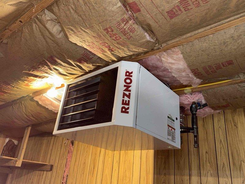 Performed a fire-safety inspection of the garage Reznor unit heater following recent attic insulation work, checking the heater, gas piping and shutoff, flexible vent connection, and galvanized flue/vent pipe for proximity to combustibles. Observed fiberglass batt insulation with torn or sagging kraft facing in the surrounding ceiling cavity and loose, displaced insulation near the vent elbow, and documented all conditions with photos.