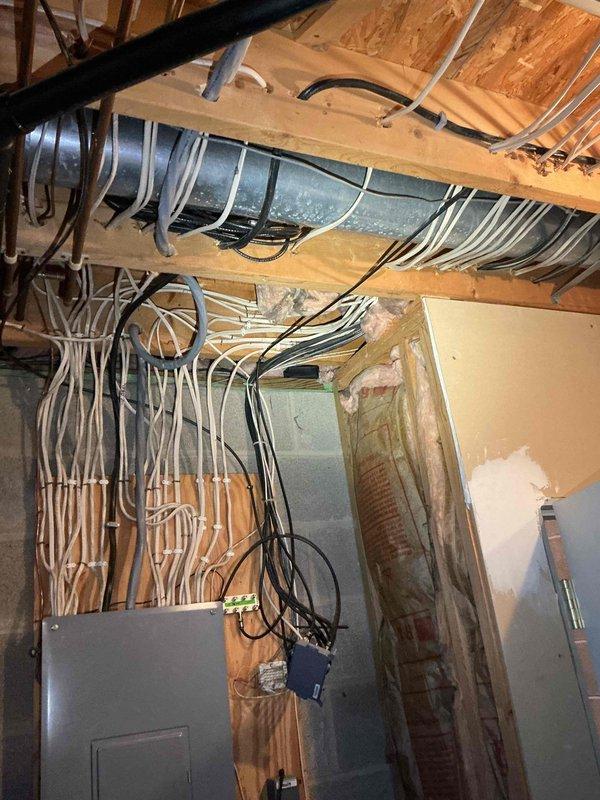 Performed an on-site evaluation for a new home generator installation, with no existing generator or related equipment present. Inspected the main electrical panel and surrounding service area in the unfinished basement, noting dense low-voltage and branch cabling routed through the ceiling joists and along the plywood panel backing, along with nearby ductwork, piping, and exposed wall cavity insulation that will need to be considered for conduit routing and transfer switch placement. Documented existing conditions with photos to support estimate preparation.