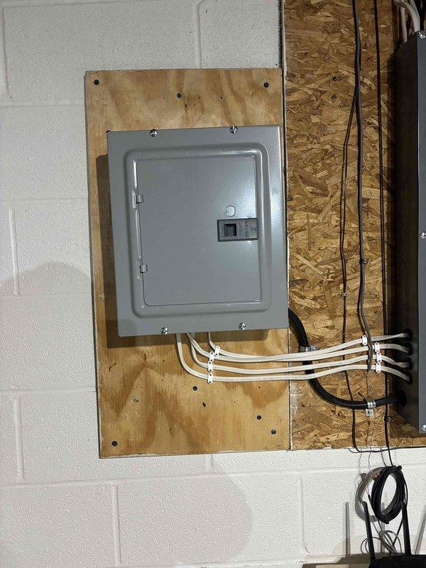Re-secured the loose plywood backing board supporting the electrical breaker panel and surge protector to the concrete block wall using appropriate masonry fasteners. Inspected the panel mounting, cable routing, and conduit connections to confirm all components were firmly supported and in good condition with no visible damage.