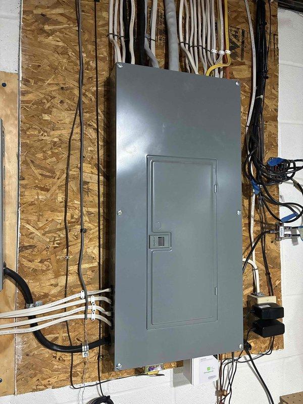 Re-secured the loose plywood backing board supporting the electrical breaker panel and surge protector to the concrete block wall using appropriate masonry fasteners. Inspected the panel mounting, cable routing, and conduit connections to confirm all components were firmly supported and in good condition with no visible damage.