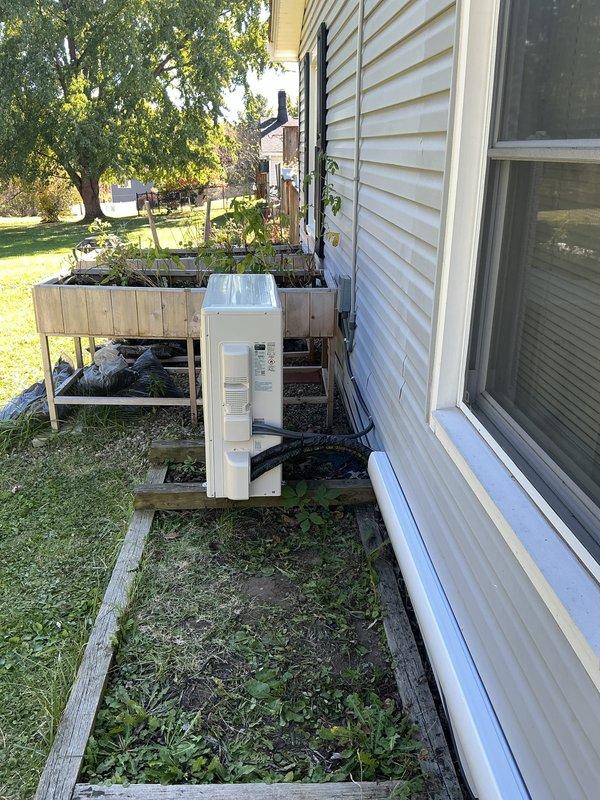 Installed a single-zone ductless mini-split system, mounting the outdoor condenser on a level timber platform at grade and the wall-mounted indoor air handler high on the interior wall. The line set was run through the exterior wall in conduit with insulated refrigerant lines and power/communication cable, and the equipment was left in clean, undamaged condition with no visible frost, rust, or defects.