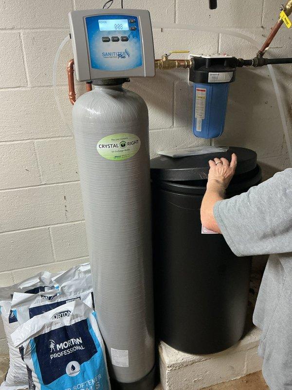 Installed a new whole-house water softener system consisting of a fiberglass resin tank with digital control head, a separate brine tank, and a cartridge filter housing, all plumbed with copper piping, brass fittings, and a bypass valve. Connected the drain and brine lines, loaded the brine tank with water softener salt, and programmed the controller. Verified all connections were secure with no leaks present.