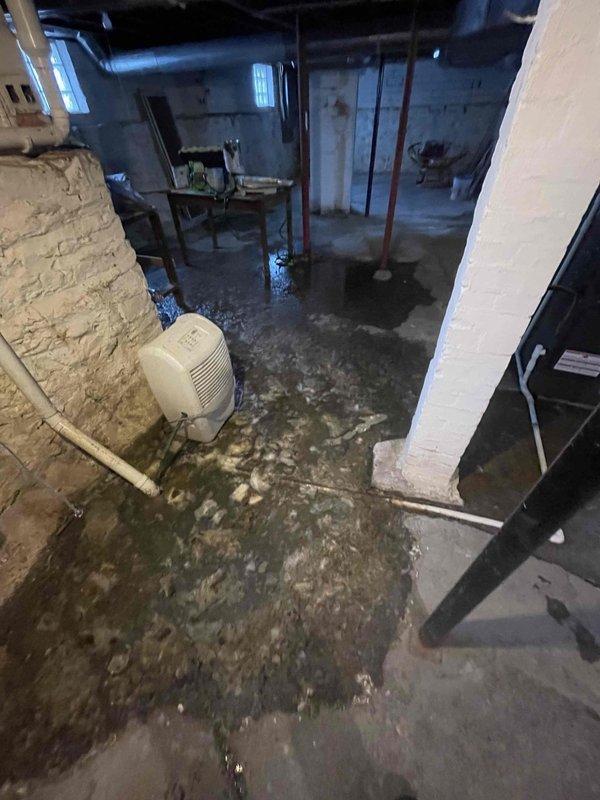 Responded to service call for backed-up sewer line reported by homeowner Kathleen Hussey. Inspection revealed significant water damage in basement with standing water on concrete floor. Located severely clogged main drain connection with substantial buildup of debris, hair, and organic material around pipe fittings, which was restricting proper water flow. Successfully cleared blockage to restore proper drainage function.