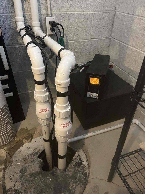 Inspected sump system and identified primary pump failure, with only backup unit operational. Observed standing water in pit with concrete deterioration and visible cracks. System includes dual PVC discharge pipes with check valves and electronic control unit for backup operation.