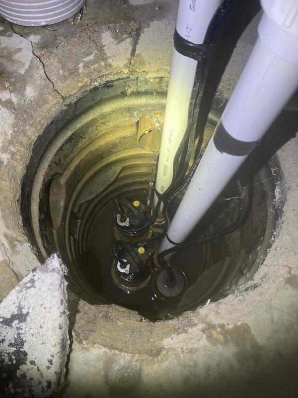 Inspected sump system and identified primary pump failure, with only backup unit operational. Observed standing water in pit with concrete deterioration and visible cracks. System includes dual PVC discharge pipes with check valves and electronic control unit for backup operation.