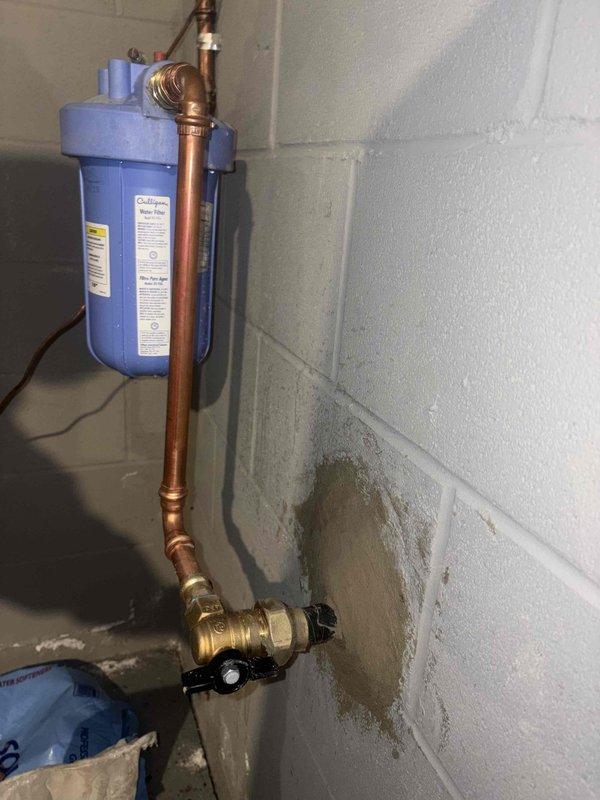 Excavated exterior area to access leaking water line. Located and repaired damaged section of pipe causing visible water spray from ground. Installed new copper piping with brass shutoff valve and connected to existing water filtration system mounted on concrete block wall.