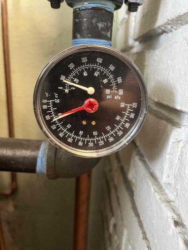 Investigated reported leak in floor suspected to be from radiant floor heating system. Inspection revealed damaged drywall with visible water damage and deterioration. Pressure gauge reading showed system pressure at approximately 60 PSI/400 kPa, which is within normal operating range, suggesting leak may be located elsewhere in the distribution system rather than in the pressure components.