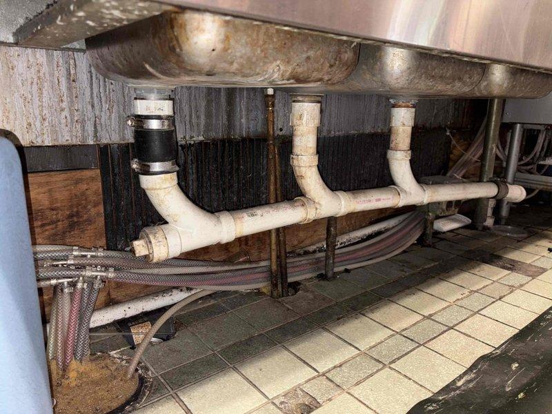 Completed inspection of commercial kitchen plumbing system, documenting significant deterioration including rust damage on stainless steel double basin sink, compromised PVC drainage assembly with visible moisture damage to surrounding wall materials, and evidence of persistent leakage. Noted ceiling infrastructure issues in kitchen area including exposed fluorescent fixture and extensive pipe corrosion. Recommended complete fixture replacement and drainage system overhaul to address water damage and prevent further structural deterioration.