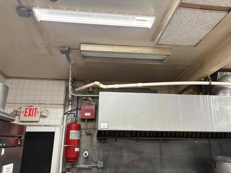 Completed inspection of commercial kitchen plumbing system, documenting significant deterioration including rust damage on stainless steel double basin sink, compromised PVC drainage assembly with visible moisture damage to surrounding wall materials, and evidence of persistent leakage. Noted ceiling infrastructure issues in kitchen area including exposed fluorescent fixture and extensive pipe corrosion. Recommended complete fixture replacement and drainage system overhaul to address water damage and prevent further structural deterioration.