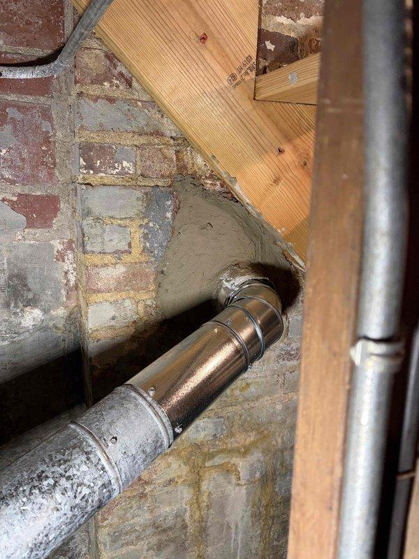 Assisted chimney technician with interior furnace vent connection through brick wall. Inspected metal ductwork and horizontal pipe connections where they penetrate the masonry wall in basement utility area. Documented existing installation with photos showing proper connection of heating unit venting system.