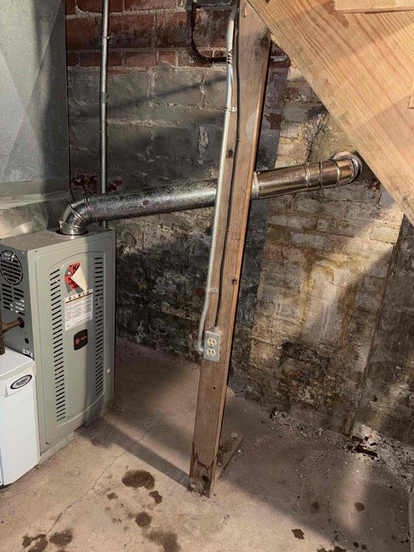 Assisted chimney technician with interior furnace vent connection through brick wall. Inspected metal ductwork and horizontal pipe connections where they penetrate the masonry wall in basement utility area. Documented existing installation with photos showing proper connection of heating unit venting system.