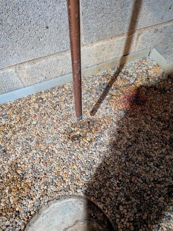 During club maintenance visit, identified corrosion on several fittings and a leaking pressure relief pipe on boiler system installed in 2024. Documented issues with photographs showing oxidation on copper piping and possible water damage to surrounding area as evidenced by discoloration in the gravel bed beneath plumbing connections.