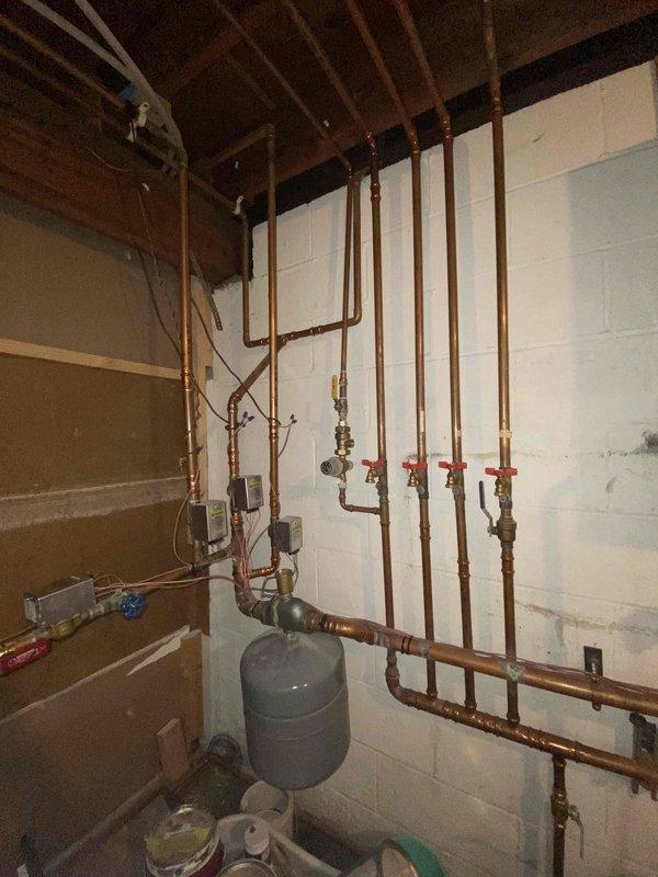 Completed estimate for combi boiler system replacement in residential basement utility room. Assessed existing copper piping infrastructure including four vertical supply lines with red-handled shut-off valves, expansion tank, and multiple horizontal distribution lines mounted to ceiling joists and concrete block walls. Evaluated current plumbing configuration, pipe routing, and connection points to determine scope of work for new combination boiler installation and integration with existing hydronic heating system.