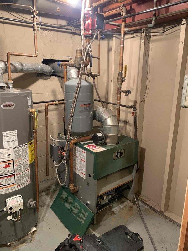 Responded to service call for 10-year-old boiler producing percolating noise. Performed comprehensive tune-up and maintenance service on residential boiler system. Conducted visual inspection of boiler unit, expansion tank, associated piping, and connections in basement mechanical room. Evaluated system operation and assessed noise complaint. Completed standard maintenance procedures including system flush to address sediment buildup causing percolating sounds. Verified proper operation of water heater and HVAC equipment in utility area. Documented equipment condition with photographs showing green boiler unit, expansion tank mounting, copper piping configuration, and overall mechanical room layout. System restored to normal operating parameters with noise issue resolved.