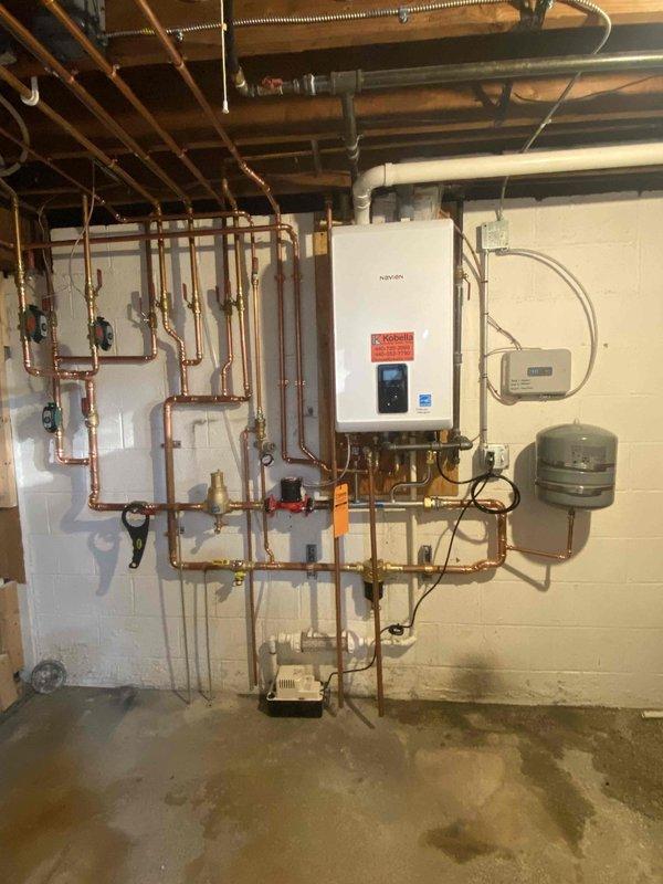 Completed installation of new wall-mounted boiler system with comprehensive hydronic piping network in basement mechanical room. Installed and configured primary heating unit with integrated manifold system featuring multiple copper supply and return lines, blue-handled isolation valves, and brass fittings for zone control. Mounted gray expansion tank and electrical control panel, ran flexible metal conduit for power supply, and completed all necessary plumbing connections including PVC venting routed through ceiling joists to exterior termination point.