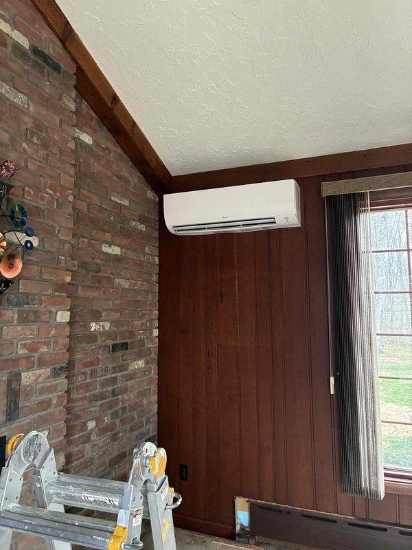 Completed ductless mini-split air conditioning system installation in residential interior space with exposed brick and wood paneling. Mounted indoor air handler unit near ceiling corner, installed exterior electrical junction box with proper leveling, and established refrigerant line connections between indoor and outdoor components. System tested and verified operational upon completion.