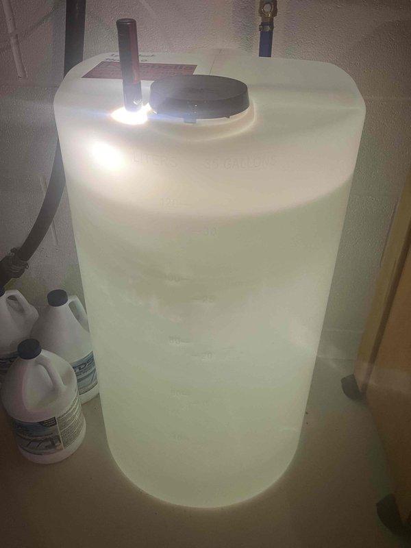 Completed installation and setup of full-scale chlorination system for water treatment. System includes white cylindrical treatment tank with dual-port configuration, blue fill line with clearly marked labeling, and associated piping connections. Chemical supply containers positioned adjacent to water heater tank for convenient access and maintenance. All components properly installed, labeled with service information, and operational markings documented for future reference.