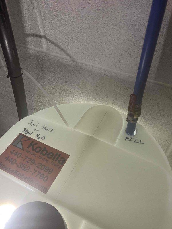 Completed installation and setup of full-scale chlorination system for water treatment. System includes white cylindrical treatment tank with dual-port configuration, blue fill line with clearly marked labeling, and associated piping connections. Chemical supply containers positioned adjacent to water heater tank for convenient access and maintenance. All components properly installed, labeled with service information, and operational markings documented for future reference.