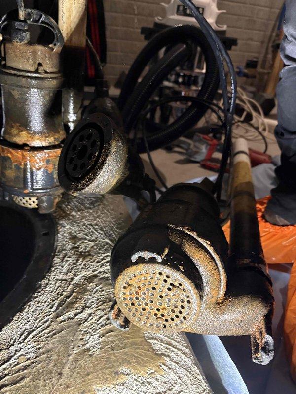 Responded to emergency service call for non-operational sump pump system in six-year-old residential property. Upon arrival, diagnosed clogged intake strainer assembly causing pump failure and preventing proper water evacuation from sump basin. Disassembled and thoroughly cleaned perforated metal strainer screen, removing accumulated debris and sediment buildup that had restricted water flow to pump mechanism. Inspected all copper and brass piping connections for integrity, noting significant condensation present on metallic surfaces indicating normal operational conditions in high-humidity basement environment. Examined battery backup system including Kobella backup battery unit housed in protective battery box with proper cable connections maintained. Verified backup power supply functionality and confirmed adequate charge levels for emergency operation during potential power outages. Reassembled strainer assembly and tested primary sump pump operation under load conditions. Confirme