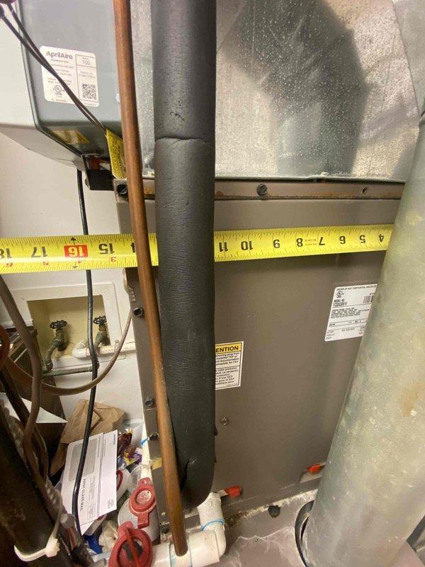 Conducted on-site assessment for HVAC system replacement in residential property. Evaluated existing furnace/air handler unit located in utility room, documenting current equipment configuration including associated ductwork, copper refrigerant lines, and gas piping connections. Measured available space constraints using tape measure to determine clearances for new equipment installation. Photographed existing gray metal furnace unit with visible manufacturer labels, surrounding ductwork infrastructure including insulated supply/return ducts, and plumbing connections with shutoff valves. Assessed mechanical room layout including vertical pipe runs, metallic ductwork transitions, and utility connections to provide accurate replacement system specifications and installation requirements for customer consideration.