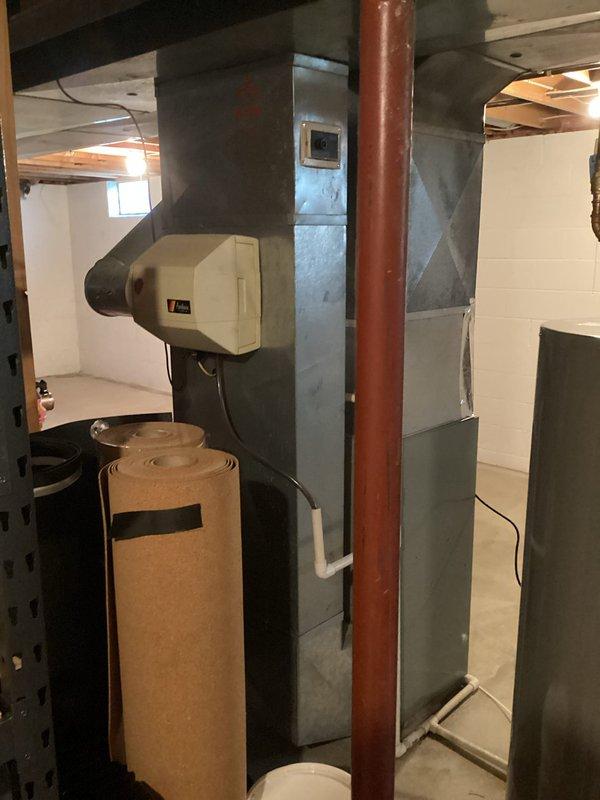 Conducted evening consultation for complete HVAC system replacement at residential property. Existing heating system experienced failure during previous winter season and requires full replacement. Evaluated current basement mechanical room configuration including existing ductwork infrastructure, water heating equipment, and humidification components. Assessed exterior access points and clearances for new equipment installation. Documented existing gray metal ductwork system with multiple bends and transitions, copper piping runs, and current humidifier unit mounted to supply plenum. Reviewed customer interest in tankless water heating option as alternative to conventional tank-style unit currently installed. Photographed basement mechanical space showing concrete block foundation walls, floor joist clearances, and utility access window for equipment removal and installation planning. Documented exterior siding conditions, deck access, and paver walkway approach for equipment delivery