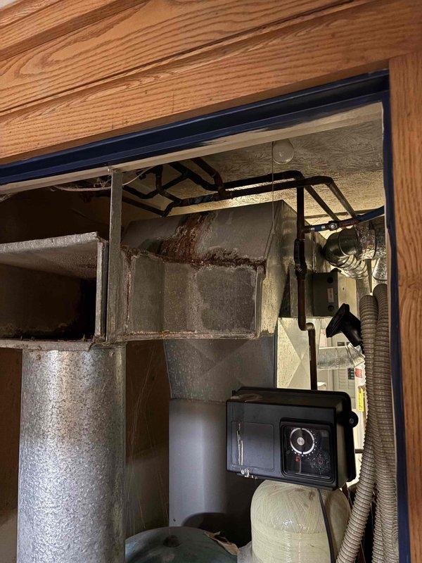 Conducted comprehensive site assessment for complete air conditioning system replacement at property with existing furnace and dual-zone ductwork infrastructure. Evaluated current HVAC configuration including aged metal ductwork showing significant dust accumulation and discoloration, existing furnace cabinet with multiple compartments, flexible and rigid duct connections, blue pressure vessel, wall-mounted air handler unit, and associated condensate drainage systems. Documented mechanical room layout and equipment condition to provide accurate replacement specifications for new AC installation that will integrate with existing forced-air heating system and dual-zone distribution network.