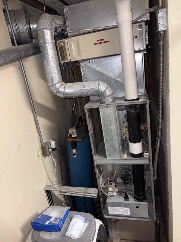 Conducted comprehensive site assessment for complete air conditioning system replacement at property with existing furnace and dual-zone ductwork infrastructure. Evaluated current HVAC configuration including aged metal ductwork showing significant dust accumulation and discoloration, existing furnace cabinet with multiple compartments, flexible and rigid duct connections, blue pressure vessel, wall-mounted air handler unit, and associated condensate drainage systems. Documented mechanical room layout and equipment condition to provide accurate replacement specifications for new AC installation that will integrate with existing forced-air heating system and dual-zone distribution network.