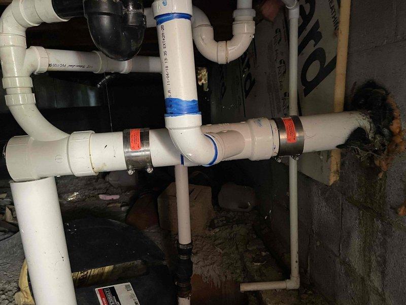 Responded to service call regarding sewer odor in bedroom potentially originating from crawlspace area. Upon inspection, discovered deteriorated pipe penetration through concrete block foundation wall with significant water damage, staining, and failed sealant material around entry point. Documented extensive moisture infiltration issues affecting wall integrity and PVC drainage system. Photographed complex sump pump discharge piping configuration showing multiple connections, elbows, and junctions with evidence of previous repairs. Identified compromised waterproofing at pipe penetration as likely source of odor infiltration into living space, with visible mold growth on foundation walls and inadequate sealing allowing sewer gases to migrate through crawlspace.