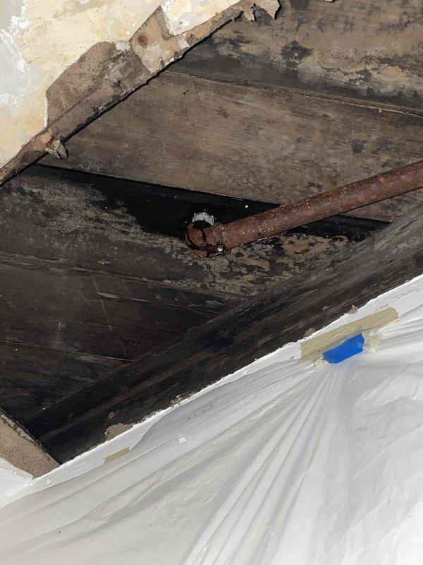 Responded to emergency call for ceiling leak originating from heating system piping. Upon arrival, found active water intrusion with extensive ceiling damage, including compromised drywall, significant water staining, and exposed structural members. Inspection revealed severely deteriorated wooden joists and beams with dark staining indicating prolonged moisture exposure and potential microbial growth. Located source of leak at corroded heating system pipe running through ceiling cavity.

Implemented immediate damage control measures by installing protective plastic sheeting to contain water flow and prevent further interior damage. Documented extensive deterioration of ceiling structure and heating pipe condition through photographic evidence. Advised customer of emergency nature of repairs needed and provided contact information for follow-up restoration work.