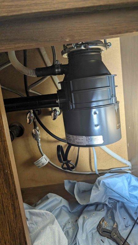 Responded to emergency call for leak under kitchen sink originating from garbage disposal. Upon arrival, inspected InSinkErator Badger 1/2 HP garbage disposal unit mounted beneath sink in fifty-year-old home. Located active leak at discharge elbow connection point on side of disposal unit. Identified worn gasket and loose mounting hardware as source of water intrusion into cabinet space. Tightened discharge tube connections, replaced deteriorated gasket, and verified proper seal at all connection points. Tested unit under running water to confirm leak was eliminated. Inspected electrical connections and power cord for safety compliance. Cleaned cabinet interior and removed standing water from base of cabinet. Unit restored to proper working condition with no further leakage detected.