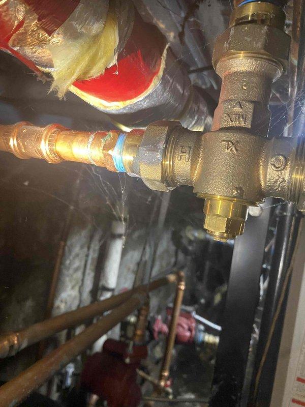 Returned to property to investigate reported leak on upstairs boiler unit. Upon inspection, identified moisture and water damage on wooden platform beneath boiler system. Located source of leak at brass T-joint valve fitting with compression-style connection. Valve showed signs of wear and minor seepage at threaded connection points. Inspected surrounding piping infrastructure, including insulated supply lines and associated ductwork. Documented conditions with photographs showing valve assembly, connection points, and water-damaged areas for service records. Recommended valve replacement and platform repair to prevent further deterioration and ensure system integrity.